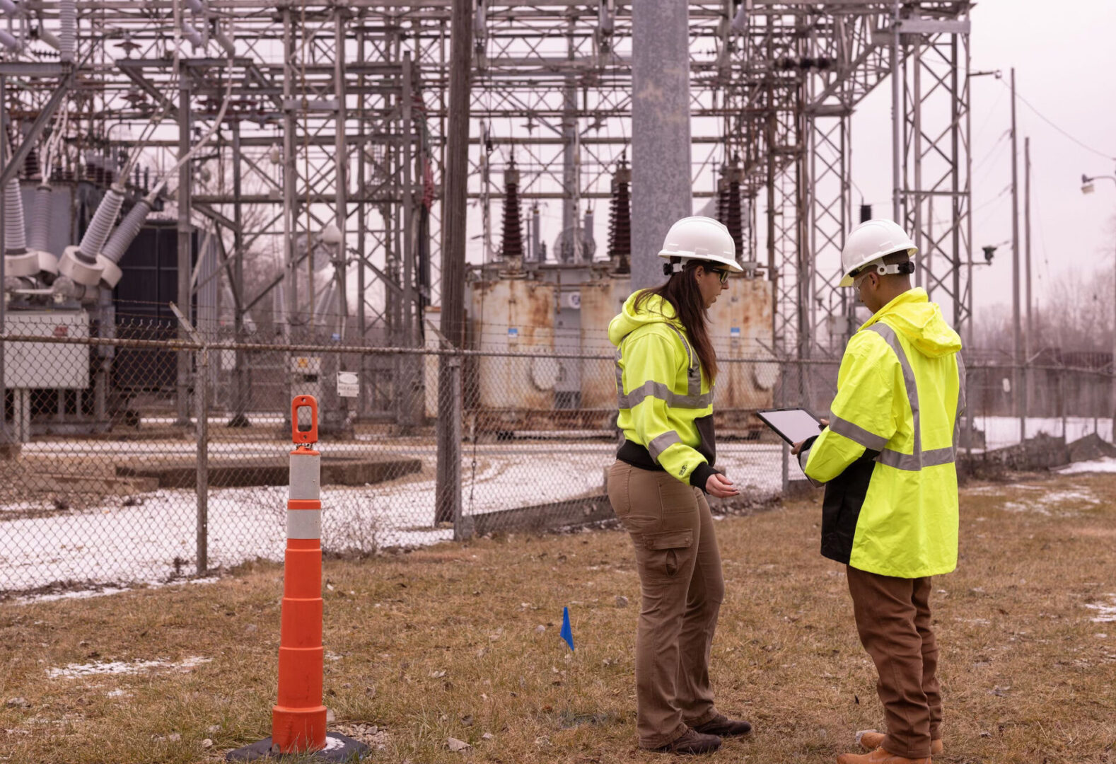 Two people in yellow jackets and hard hats standing next to a cone.