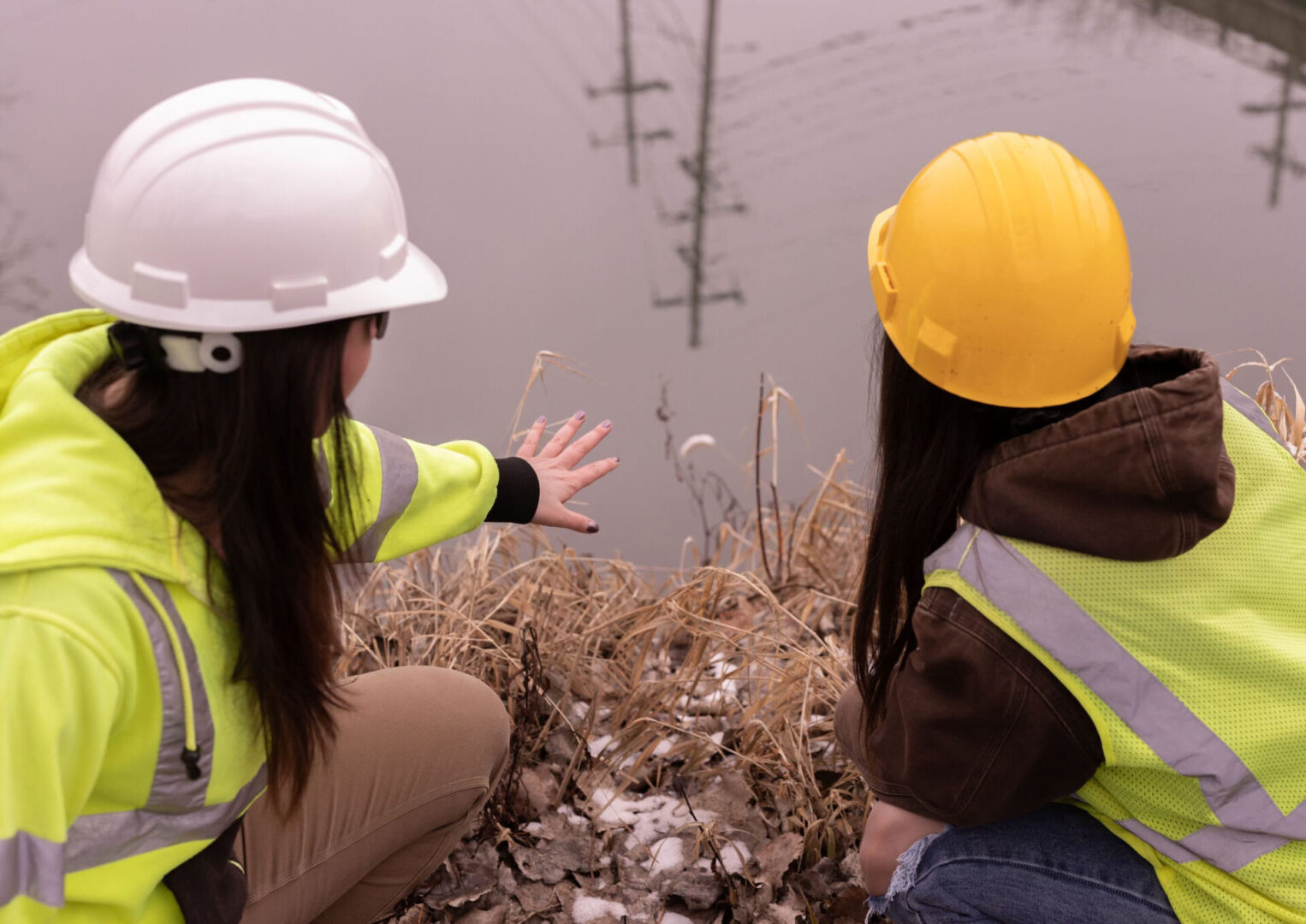 Two women in hard hats and yellow vests are looking at a pile of rubble.